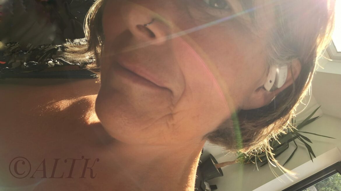Woman with short hair, we can see the headphone in one of her ears and a soft smile on her lips, a shaft of sunlight is cutting across her face